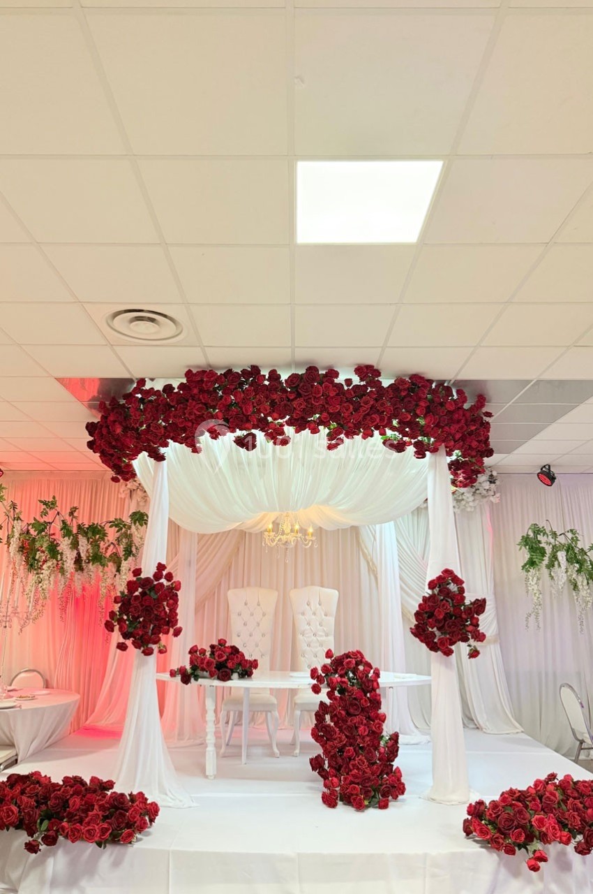 Décoration de mariage avec arche ornée de roses rouges, chaises blanches et drapés blancs dans une salle éclairée.