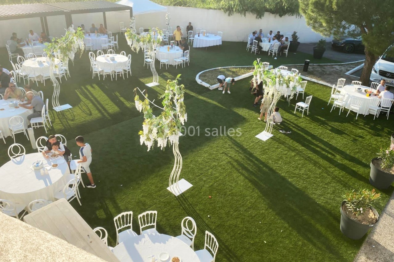 Jardin aménagé pour un événement avec des tables rondes, des chaises blanches et des décorations florales suspendues.