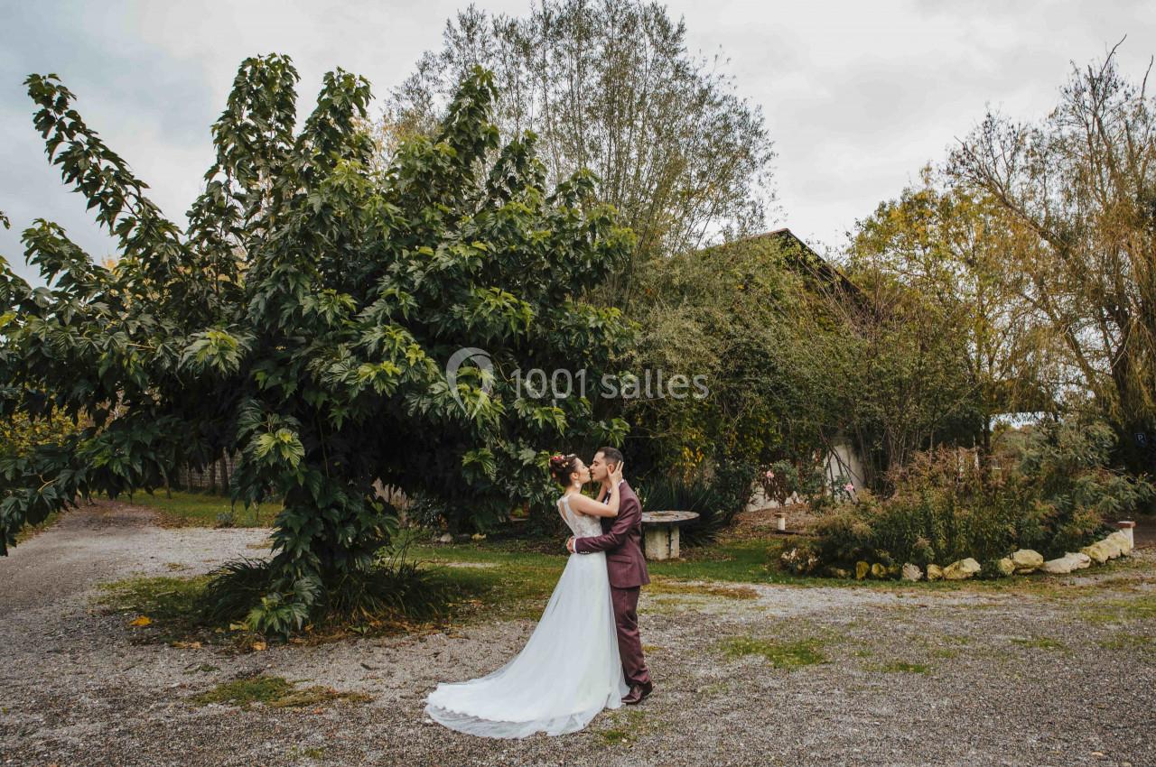 Un couple en tenue de mariage s'embrasse dans une cour extérieure bordée d'arbres et de végétation.