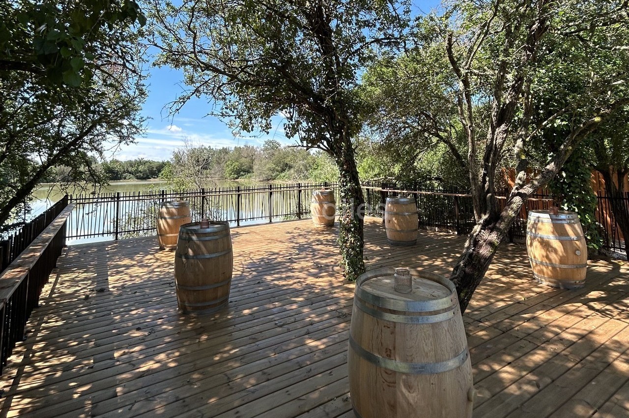 Terrasse en bois ombragée avec des tonneaux en guise de tables, surplombant un plan d'eau entouré de végétation.