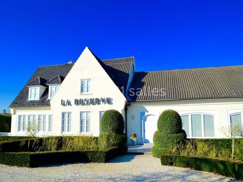 Façade d'un bâtiment blanc avec toit en ardoise, entouré de haies taillées et d'un ciel bleu dégagé.