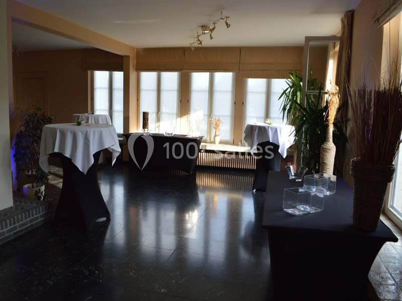 Salle lumineuse avec grandes fenêtres, tables hautes drapées de nappes blanches et décorations végétales.