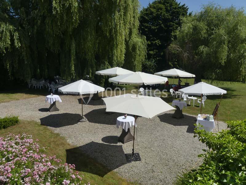 Espace extérieur avec tables et parasols blancs disposés sur du gravier, entouré de verdure et d'arbres.