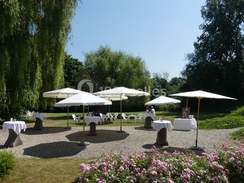 Tables avec nappes blanches et parasols disposés dans un jardin verdoyant par une journée ensoleillée.