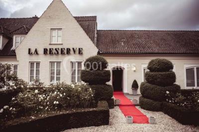 Salle de réception avec tables rondes dressées, nappes blanches, assiettes dorées et décoration florale centrale.