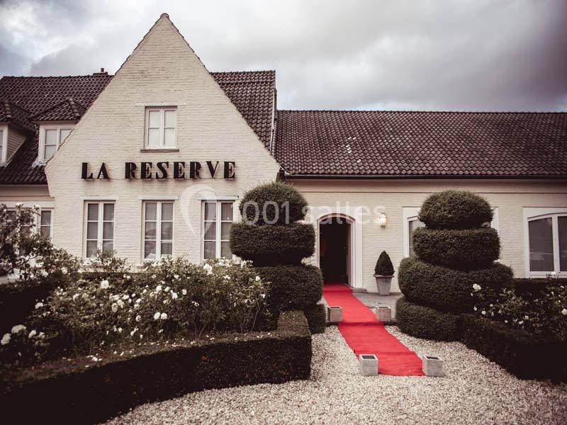 Façade d'un bâtiment blanc avec toit en tuiles, entrée ornée d'un tapis rouge et jardin taillé.