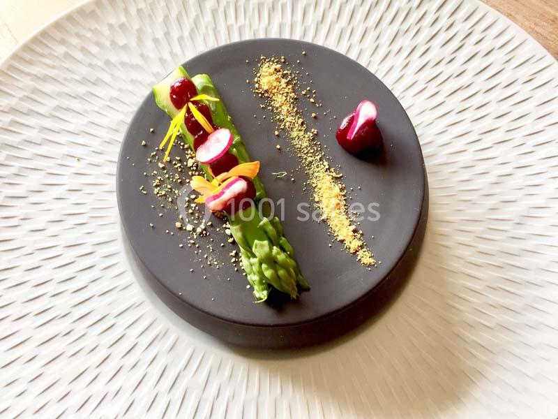 Assiette gastronomique avec asperges, cerises et poudre dorée, présentée sur une base noire et une assiette texturée.