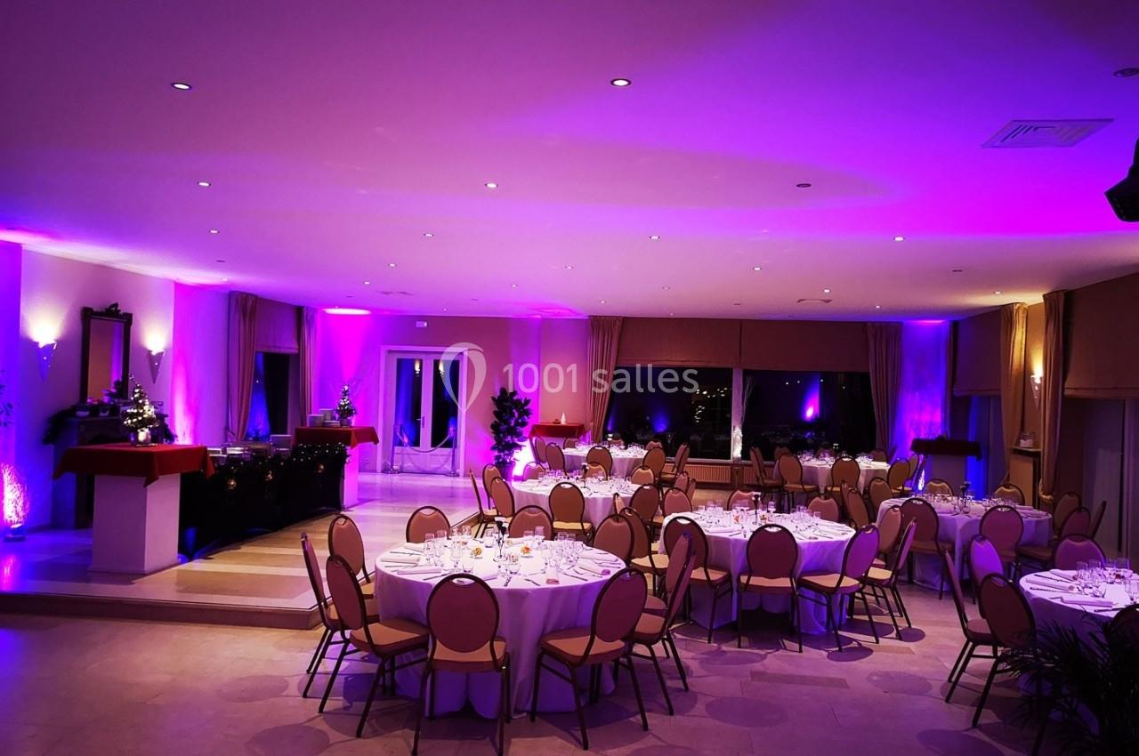 Salle de réception décorée avec des tables rondes dressées, éclairage d'ambiance violet et baies vitrées en arrière-plan.