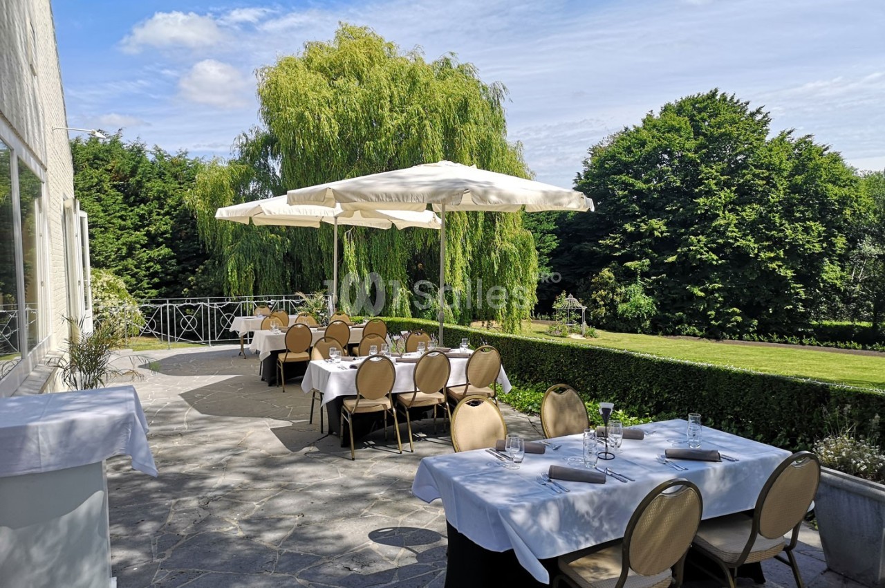 Terrasse d'un restaurant avec tables dressées, parasols blancs et vue sur un jardin arboré par une journée ensoleillée.