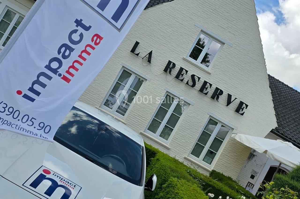 Bâtiment blanc avec l'inscription ’La Réserve’, une voiture et des drapeaux portant le logo ’Impact Immo’ au premier plan.