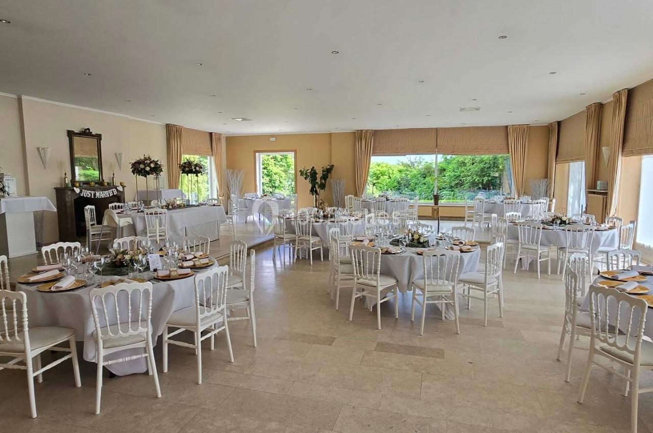 Salle lumineuse aménagée pour un événement, avec des tables rondes dressées et des chaises blanches.