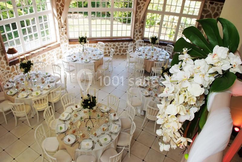Salle lumineuse décorée pour un mariage, avec tables rondes dressées, chaises blanches et orchidées blanches en premier plan.