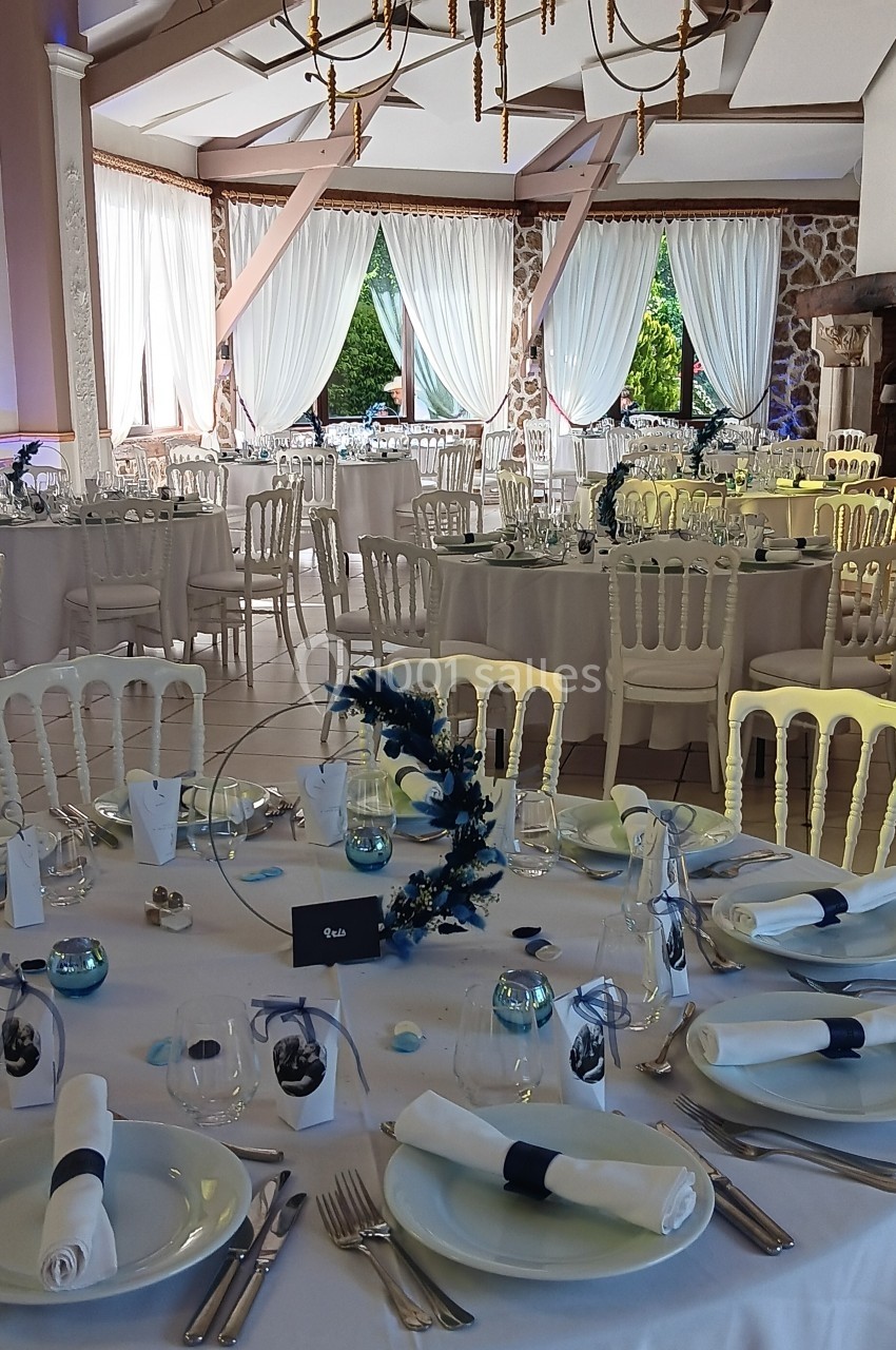 Salle de réception décorée avec des tables rondes dressées, nappes blanches et centres de table floraux bleus.