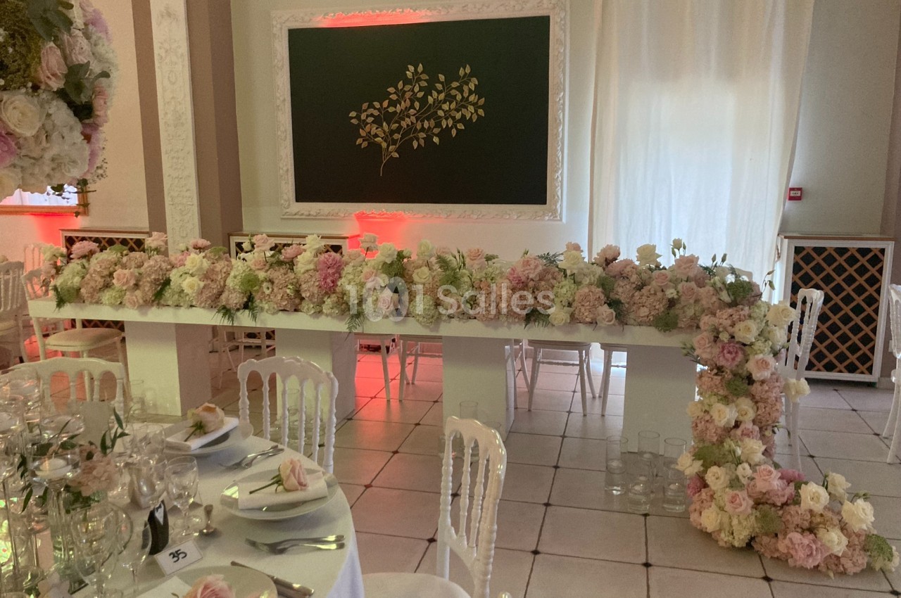 Table décorée de fleurs pastel dans une salle de réception, avec tableau noir orné d'un motif végétal en arrière-plan.