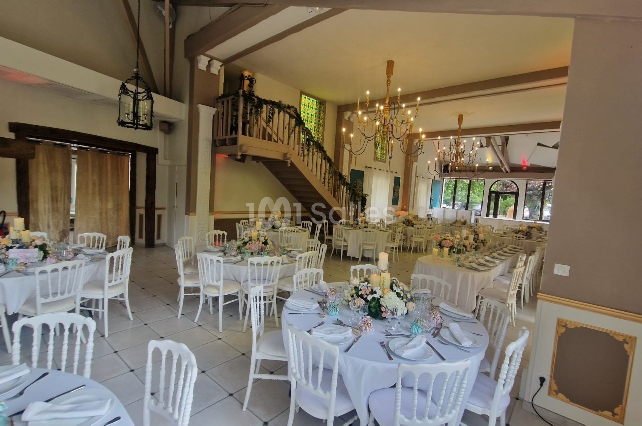 Salle de réception décorée avec des tables rondes, nappes blanches, chaises assorties et arrangements floraux.