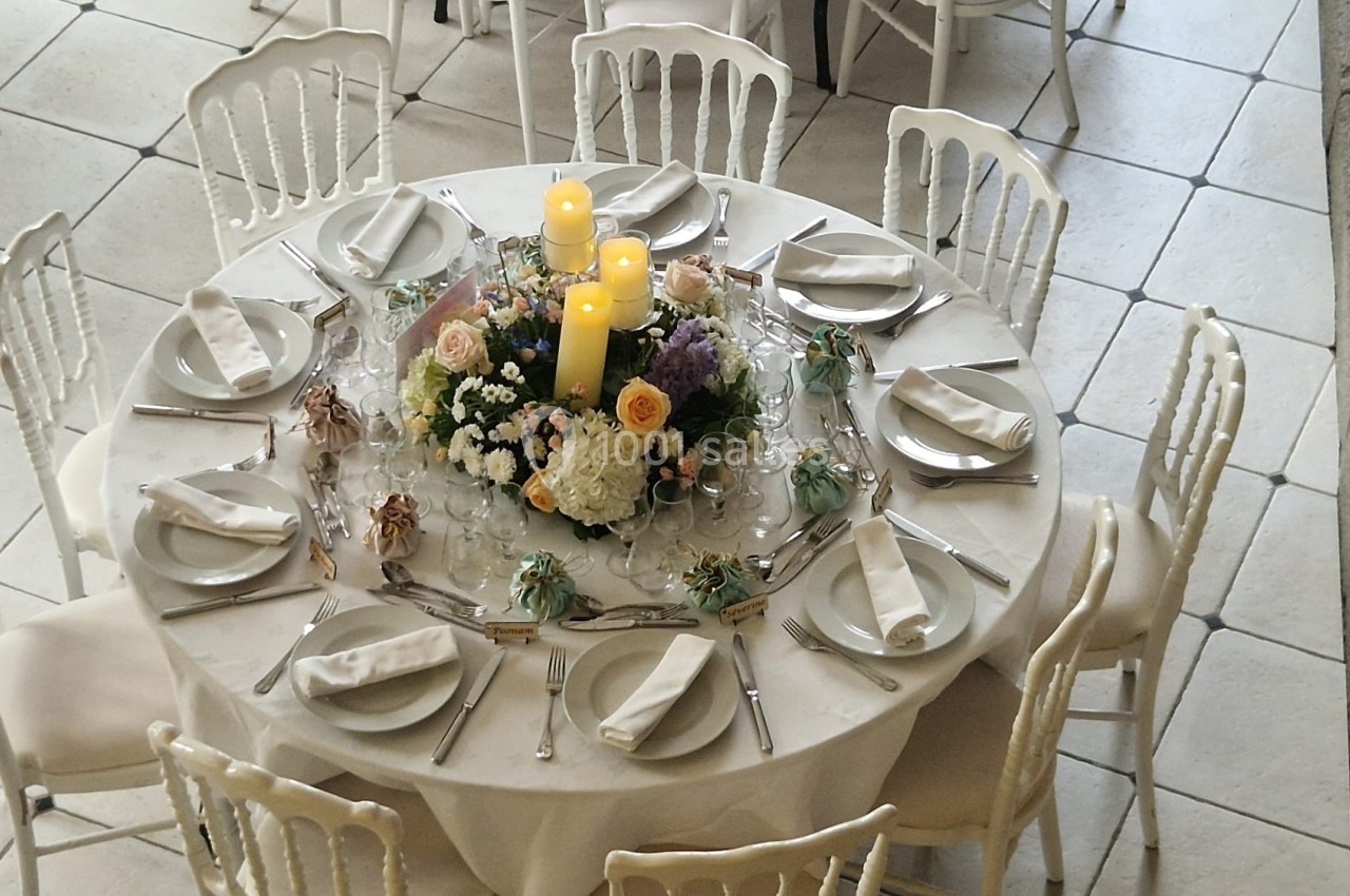Table ronde décorée pour un événement, avec bougies, fleurs et couverts disposés autour d'assiettes blanches.