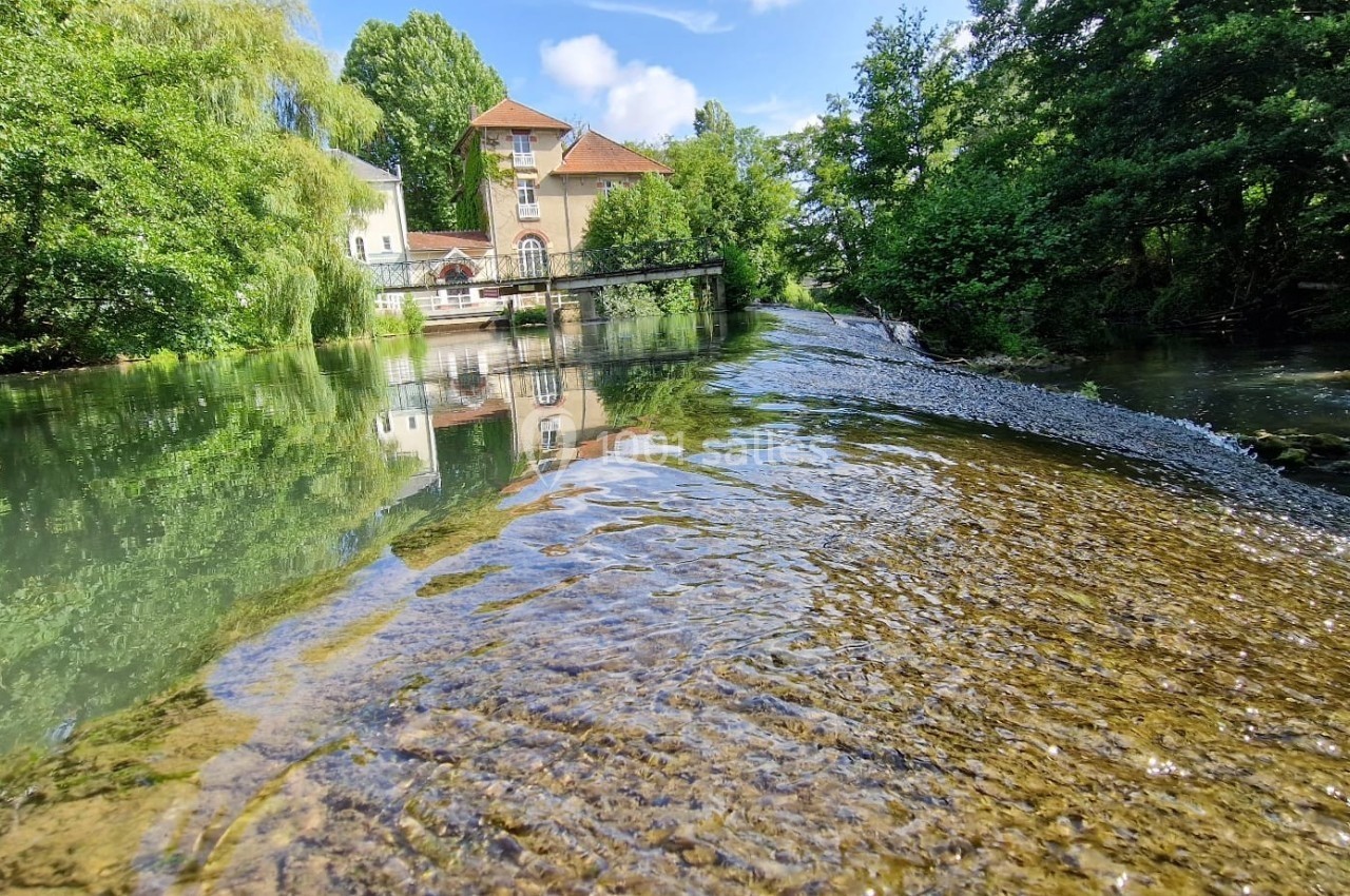 Rivière peu profonde avec un léger courant, bordée d'arbres, et un bâtiment en arrière-plan sous un ciel dégagé.