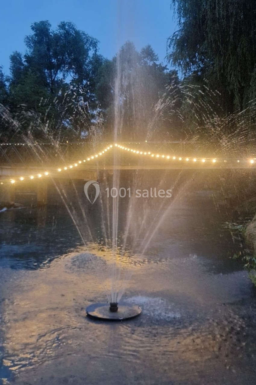 Jet d'eau illuminé au crépuscule, entouré de guirlandes lumineuses et d'arbres en arrière-plan.