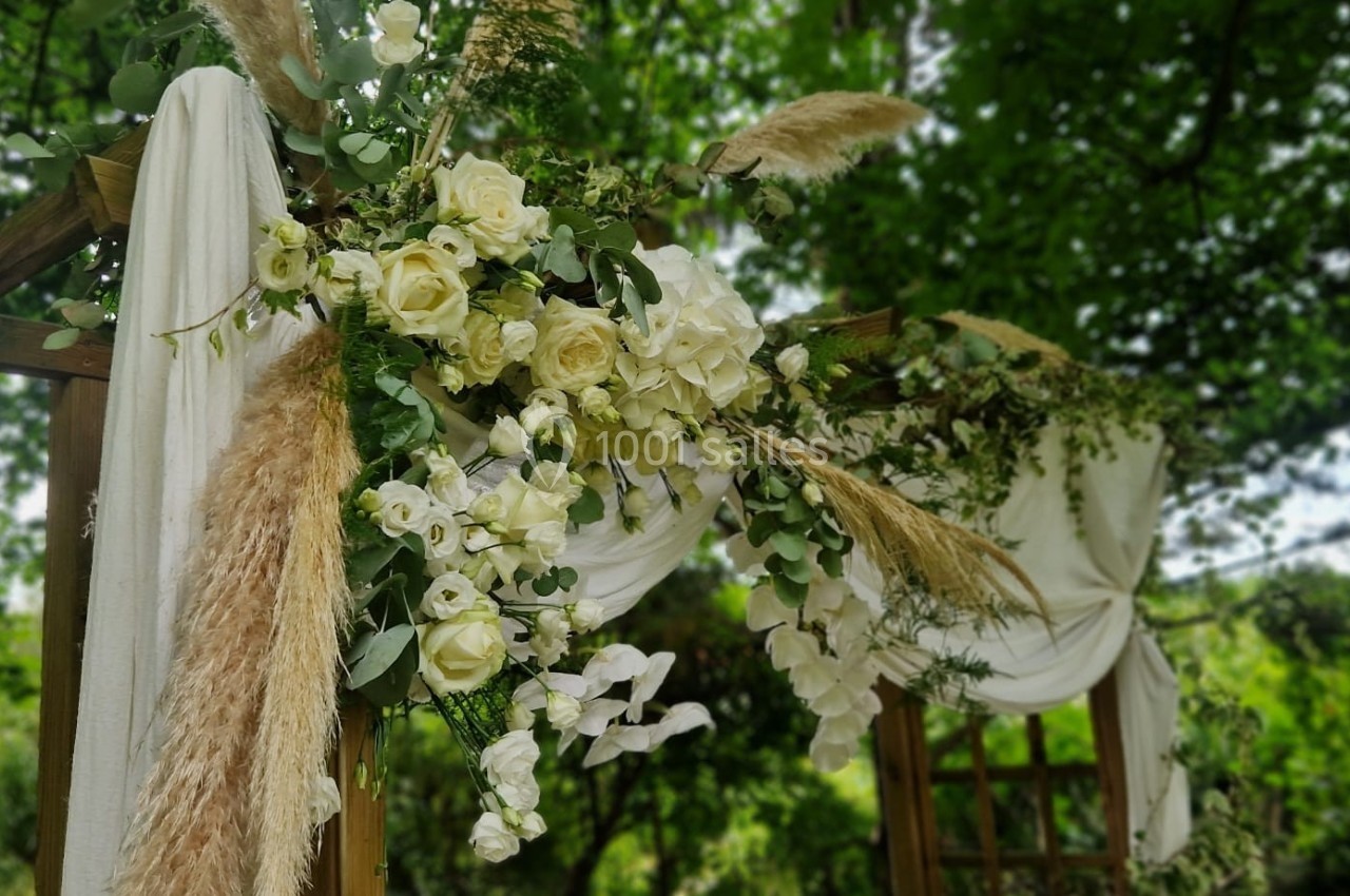 Arche en bois décorée de fleurs blanches, de feuillage et de drapés, installée dans un jardin verdoyant.