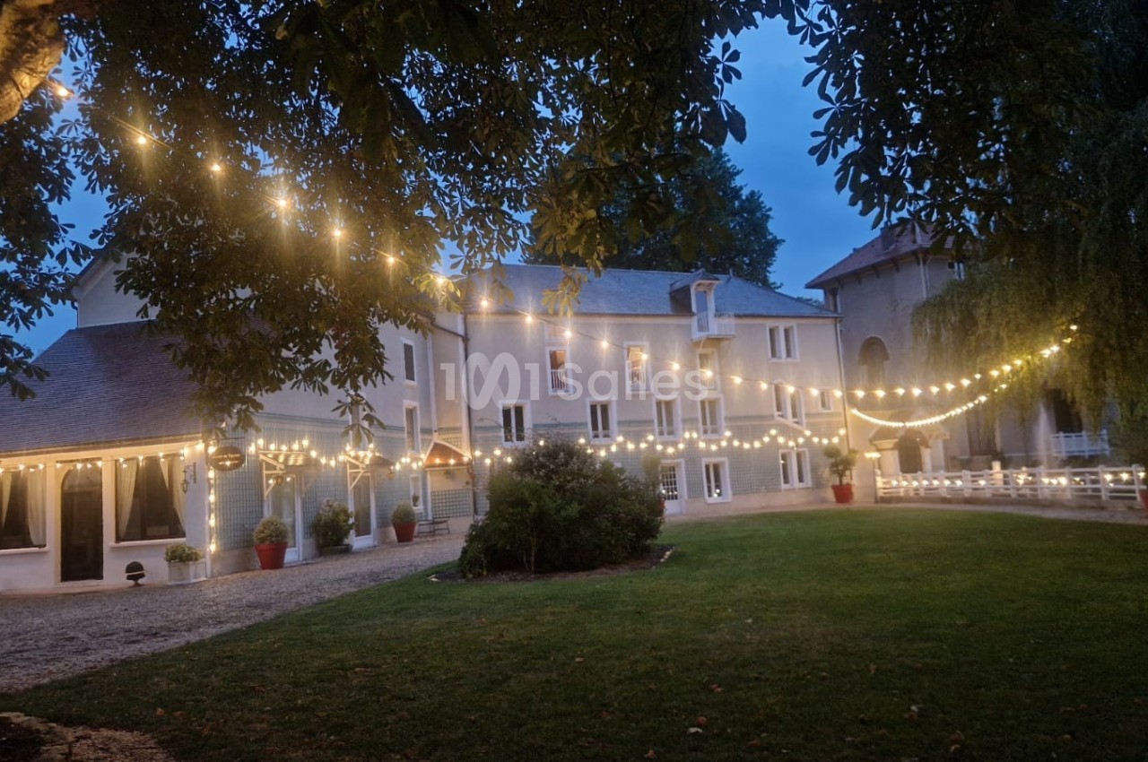 Grande maison éclairée par des guirlandes lumineuses dans un jardin en soirée, entourée d'arbres.