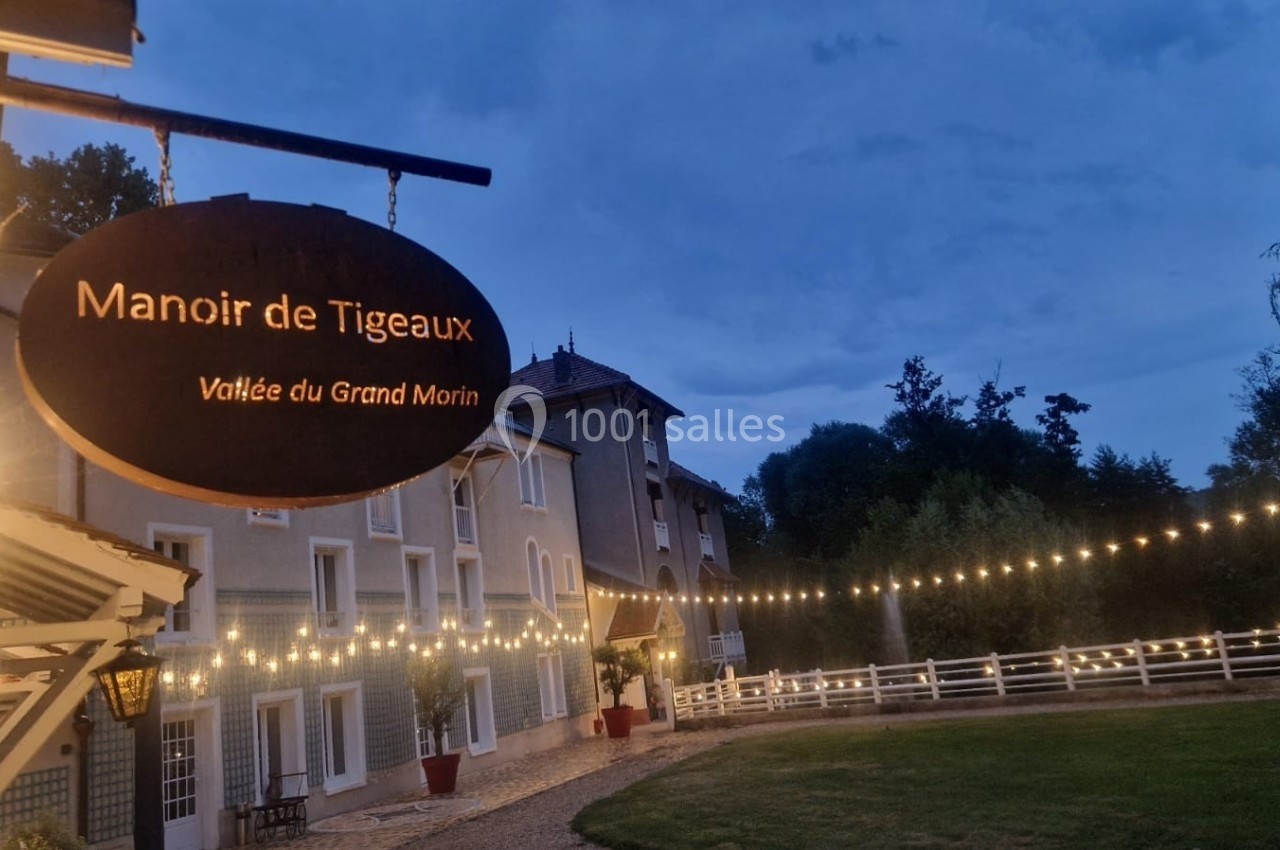 Enseigne du Manoir de Tigeaux éclairée, avec guirlandes lumineuses et bâtiment en arrière-plan au crépuscule.