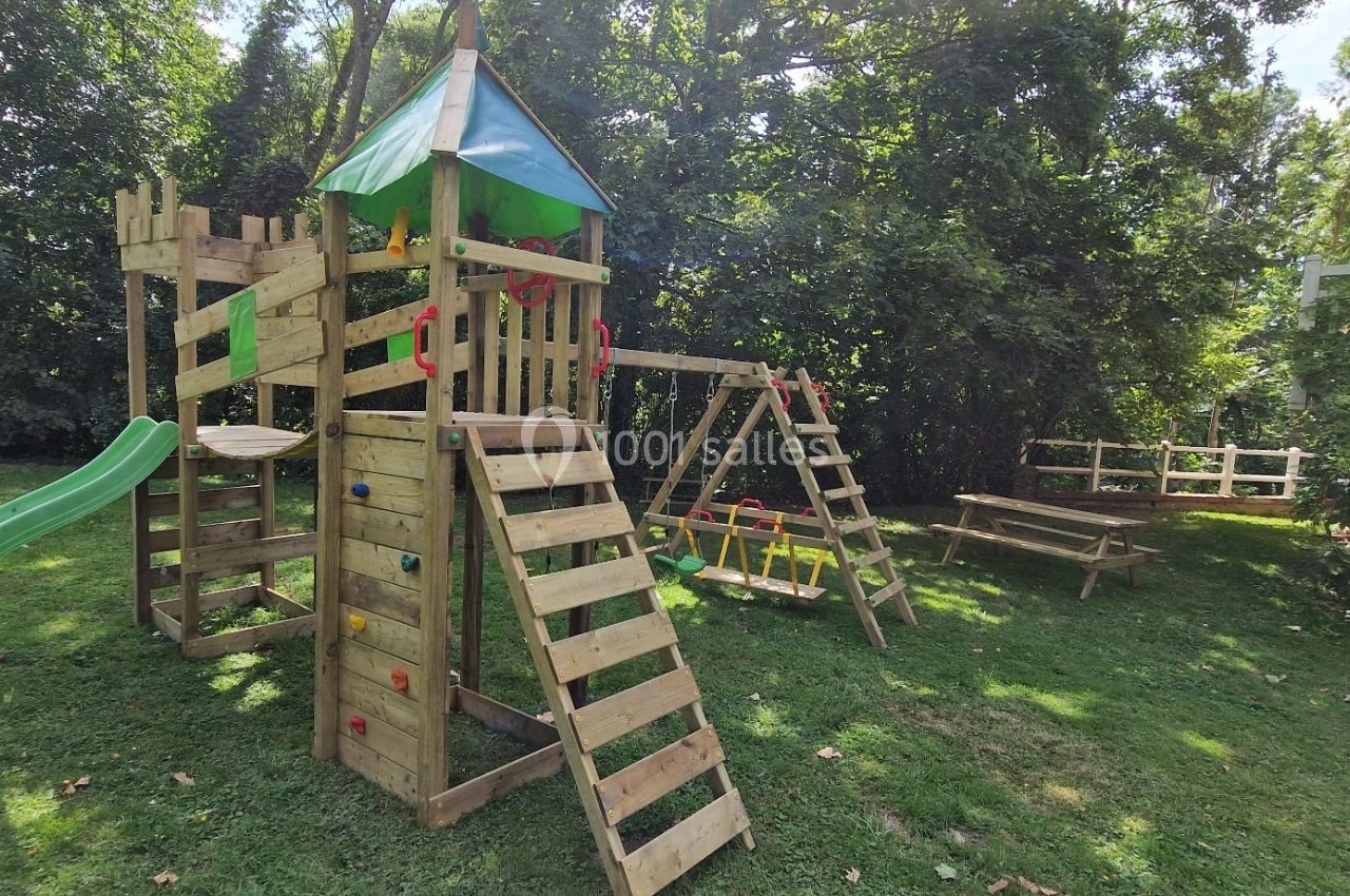 Structure de jeux en bois avec toboggan, mur d'escalade et balançoires, installée sur une pelouse entourée d'arbres.