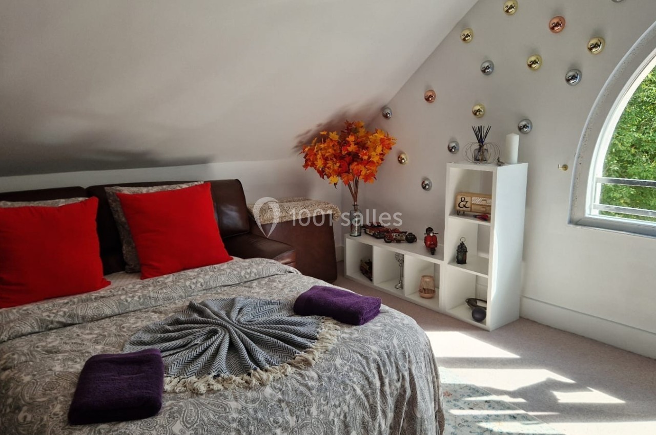 Chambre mansardée avec lit double, coussins rouges, étagère décorative, fenêtre en arche et arbre décoratif coloré.