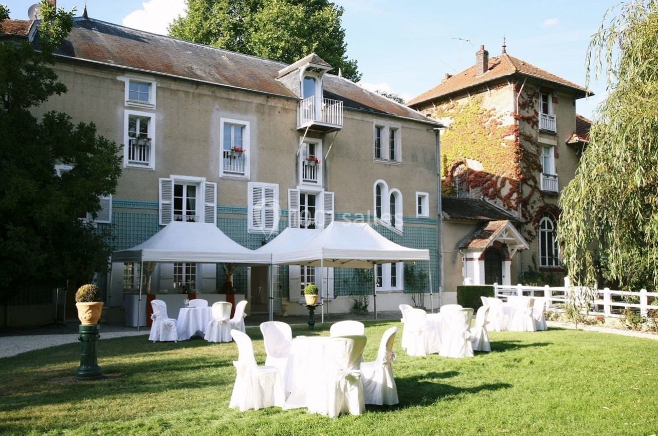 Jardin avec tables et chaises blanches sous des tonnelles, devant une grande maison ancienne entourée de verdure.