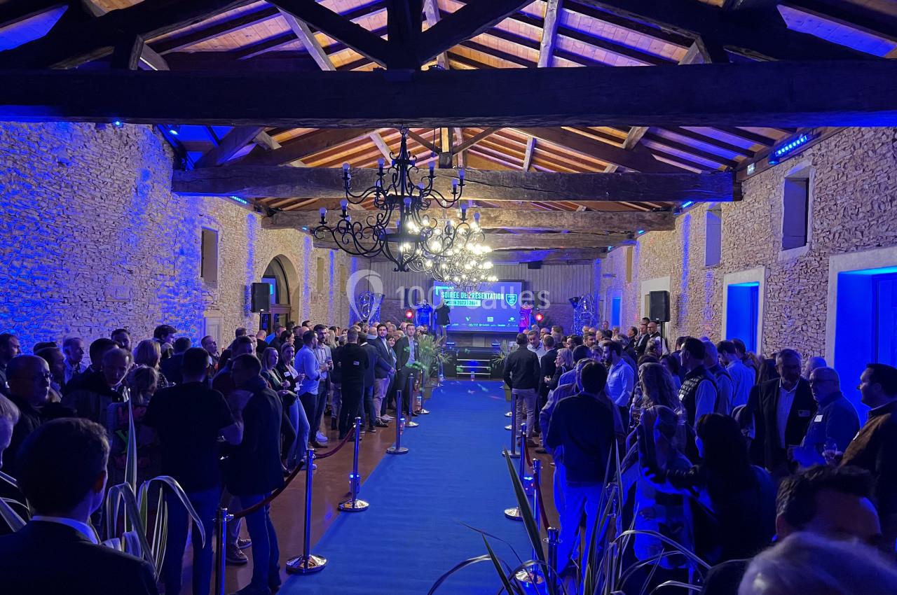 Salle en pierre avec plafond en bois, éclairée en bleu, remplie de personnes lors d'un événement avec une scène au fond.