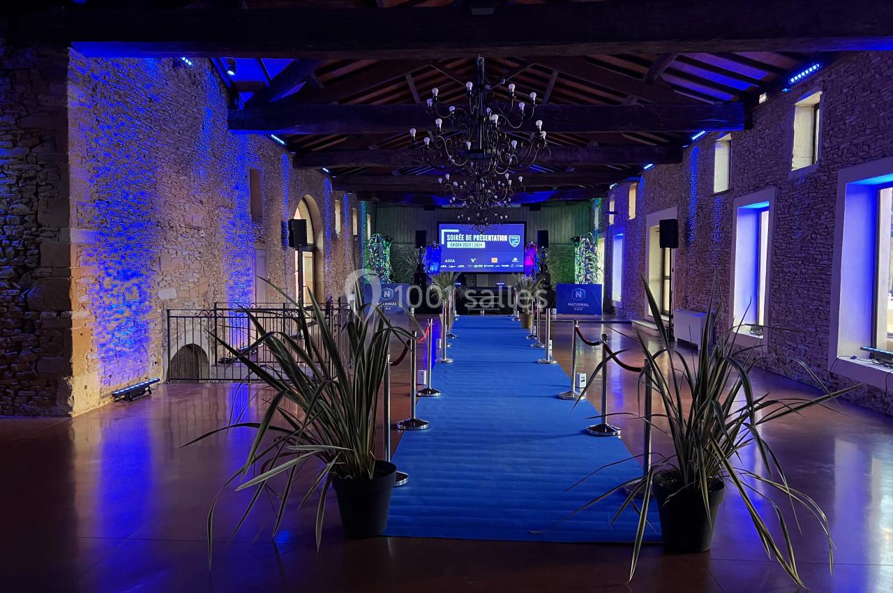 Salle en pierre avec un tapis bleu menant à une scène éclairée, décorée de plantes et de luminaires suspendus.