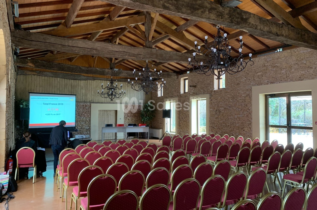 Salle de conférence avec des rangées de chaises rouges, un écran de projection allumé et des poutres apparentes au plafond.
