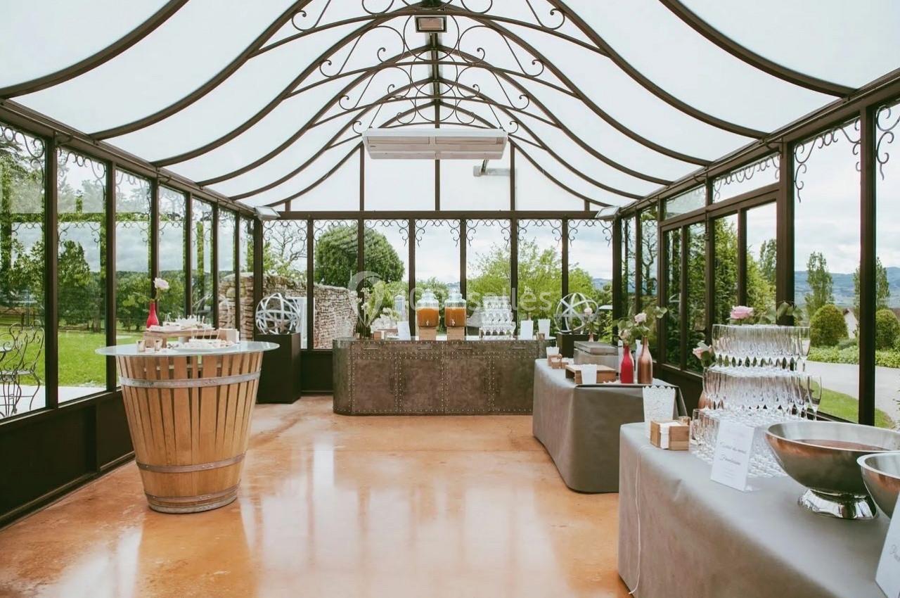 Salle lumineuse avec verrière, tables décorées pour un événement, boissons et buffet disposés sur des comptoirs.