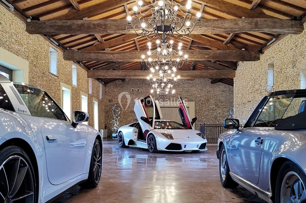 Trois voitures de sport blanches exposées dans une salle en pierre avec un plafond en bois et un grand lustre central.