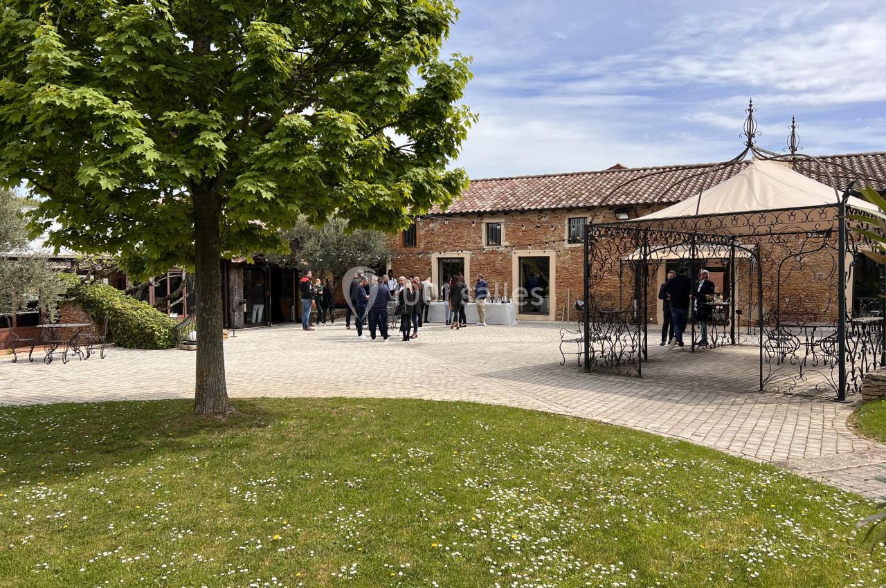 Cour pavée avec un arbre, une tonnelle en fer forgé et des personnes rassemblées devant un bâtiment en briques.