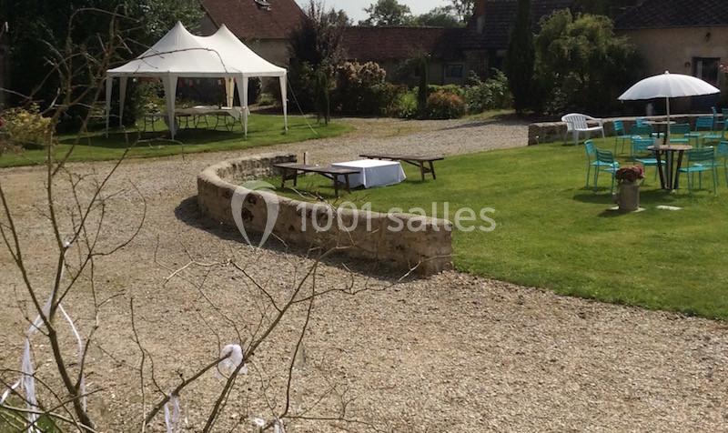 Cour extérieure avec pelouse, tables et chaises sous un parasol, et une tente blanche en arrière-plan.