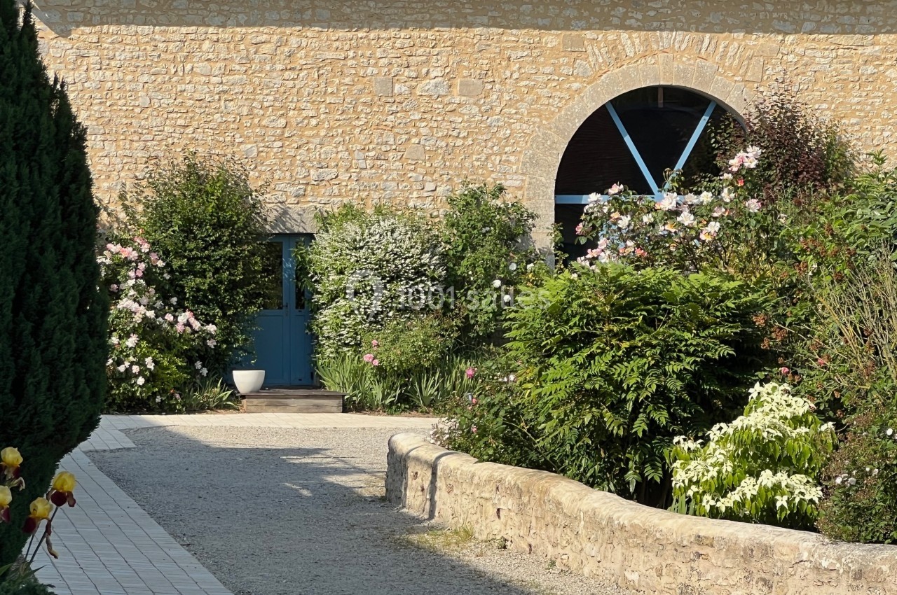 Façade en pierre avec porte bleue, entourée de buissons fleuris et d'un chemin pavé bordé d'un muret.