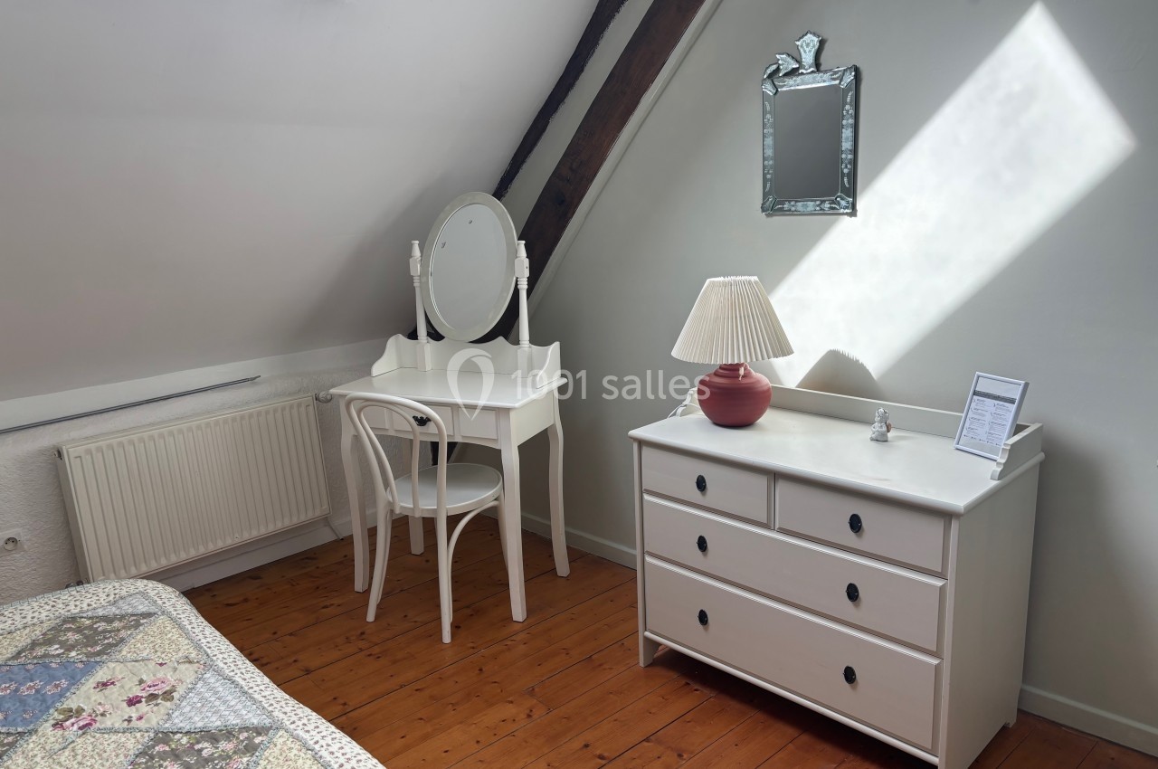 Chambre mansardée avec coiffeuse blanche, chaise, commode assortie, miroir et lumière naturelle provenant d'une fenêtre.
