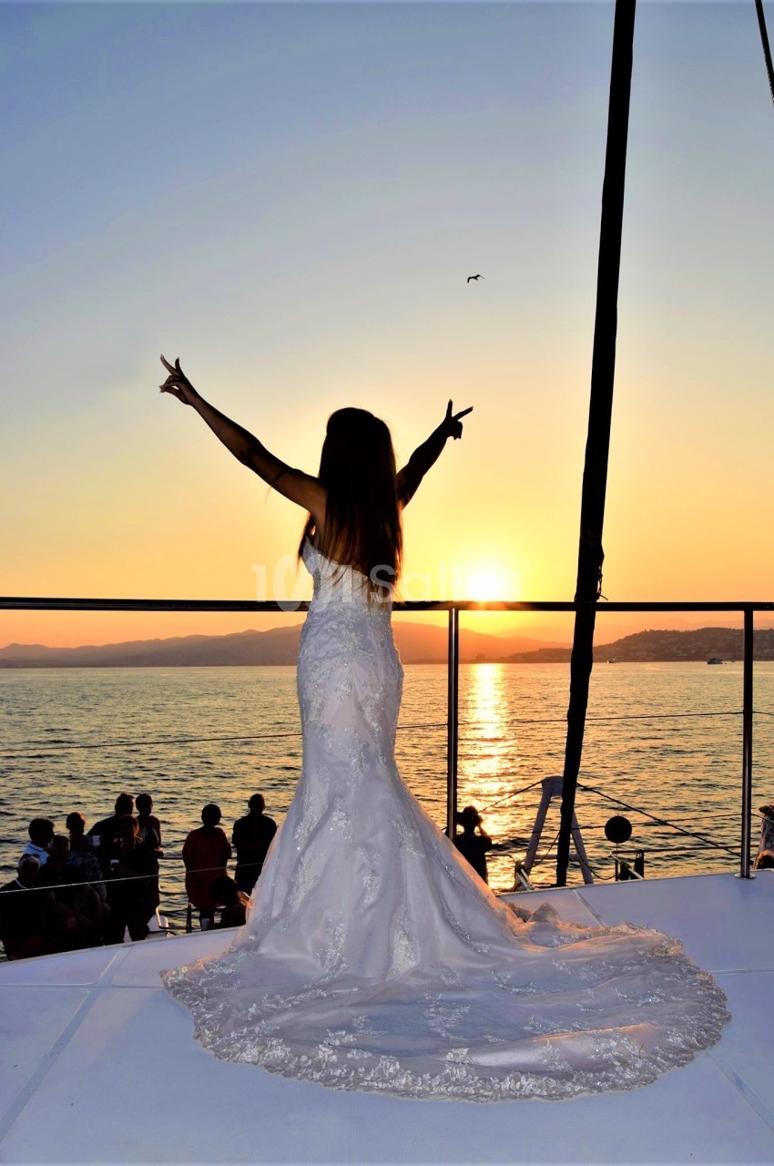 Femme en robe de mariée levant les bras face à un coucher de soleil sur un bateau, entourée de spectateurs.
