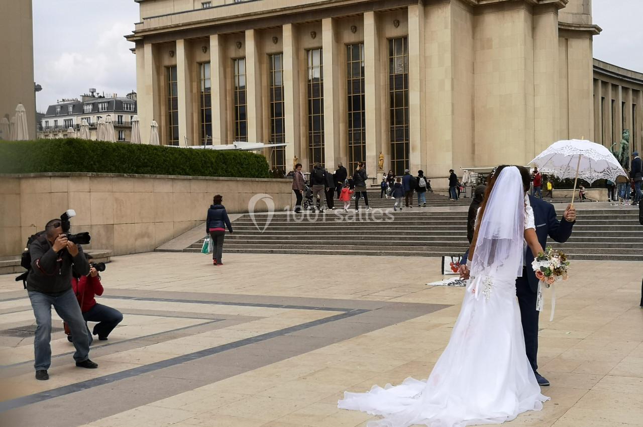 Un couple en tenue de mariage pose devant le Palais de Chaillot, entouré de passants et de photographes.