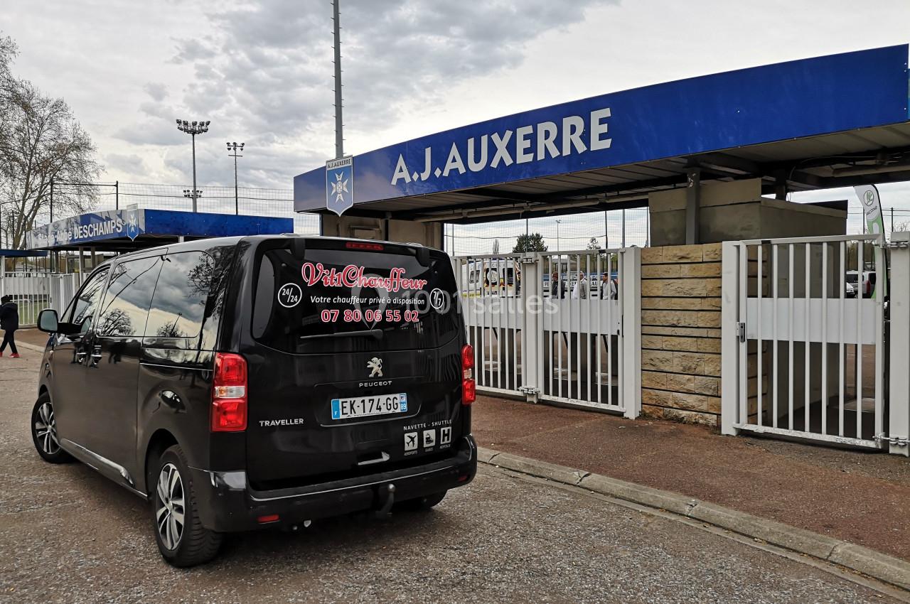 Un minivan noir stationné devant l'entrée principale du stade de l'A.J. Auxerre par temps nuageux.
