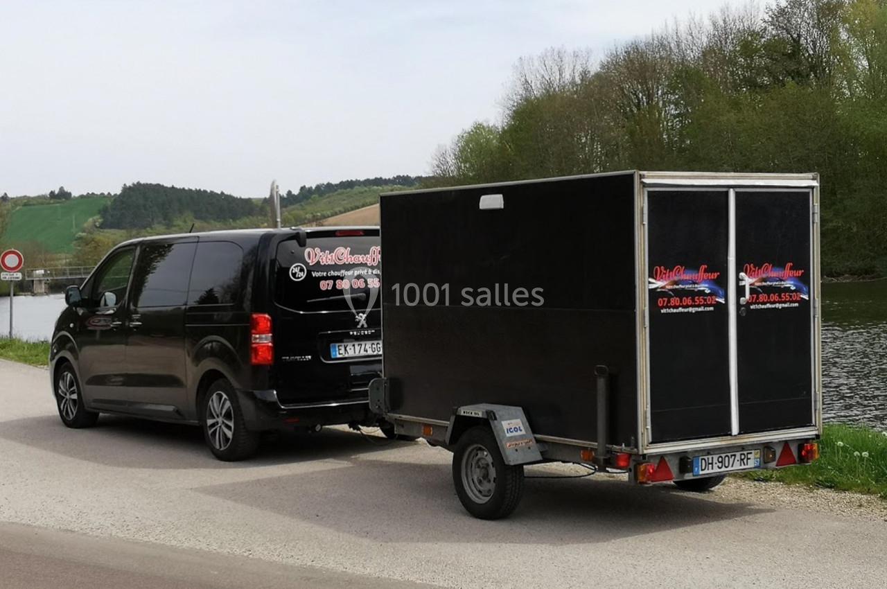 Un van noir Peugeot tractant une remorque noire sur une route près d'un cours d'eau et d'une zone boisée.