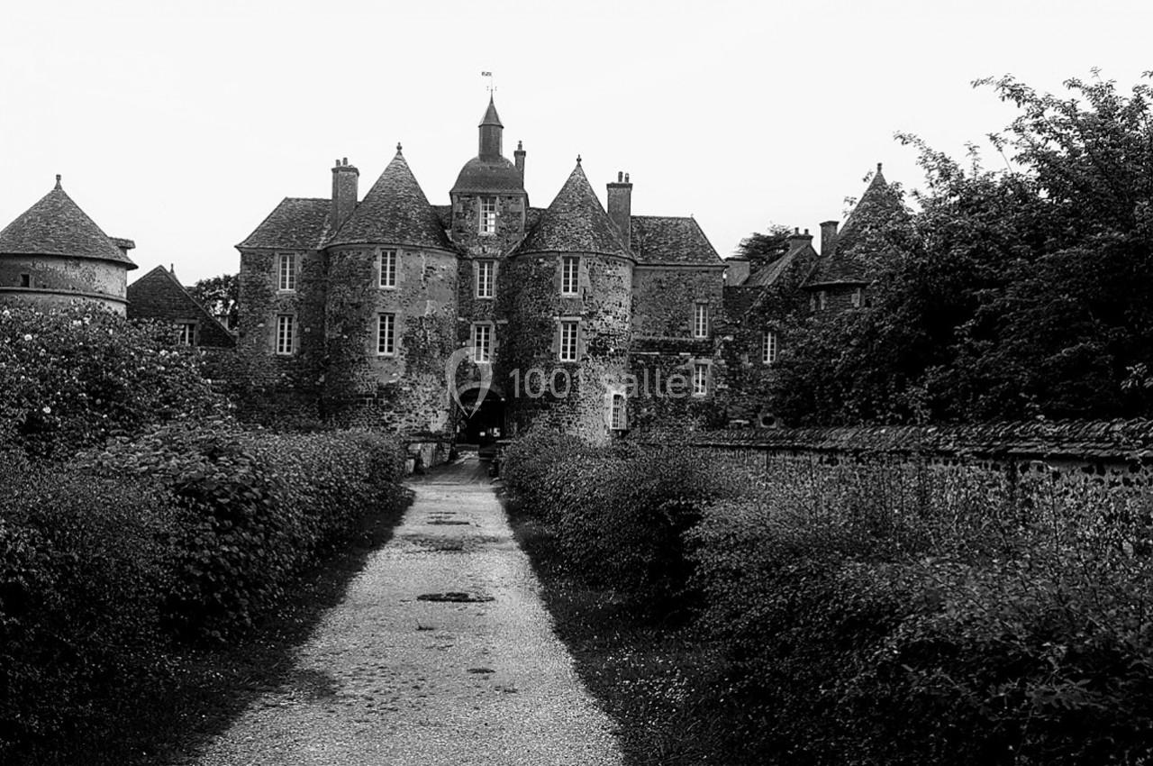 Chemin bordé de végétation menant à un château ancien en pierre avec tours et toits en ardoise.