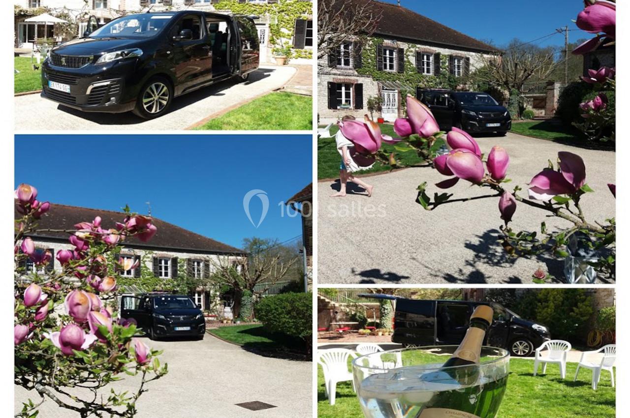 Van noir garé dans la cour d'une maison en pierre, entouré de magnolias en fleurs et de chaises blanches.