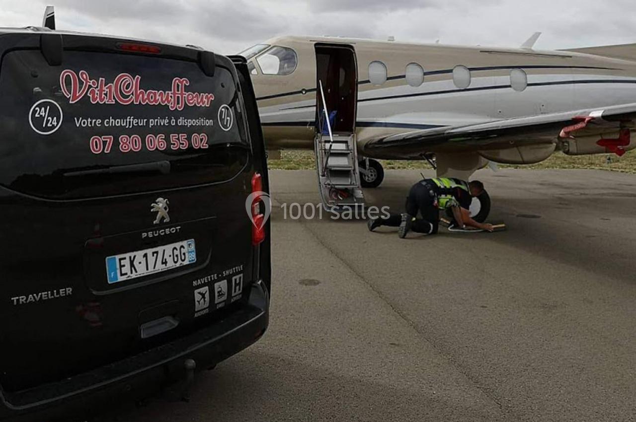 Un van noir stationné près d'un jet privé sur un tarmac, avec une personne agenouillée près de l'avion.