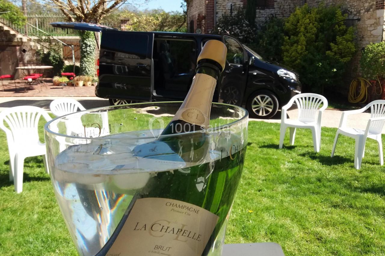 Bouteille de champagne dans un seau à glace sur une table, avec jardin, chaises blanches et véhicule en arrière-plan.