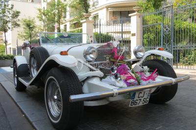 Voiture ancienne blanche décorée de fleurs roses, stationnée devant un bâtiment historique et un jardin.