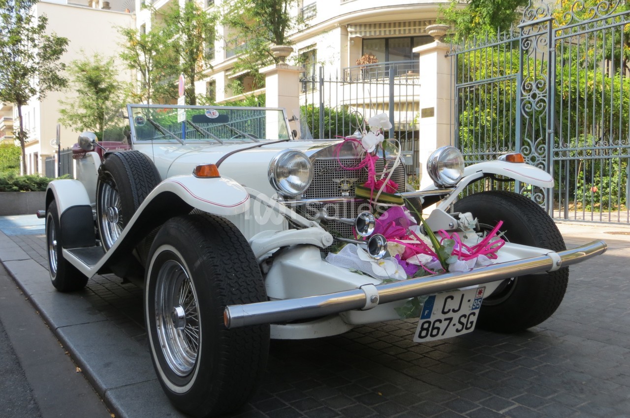 Voiture ancienne blanche décorée de rubans roses et blancs, garée devant des bâtiments résidentiels et une grille en fer…