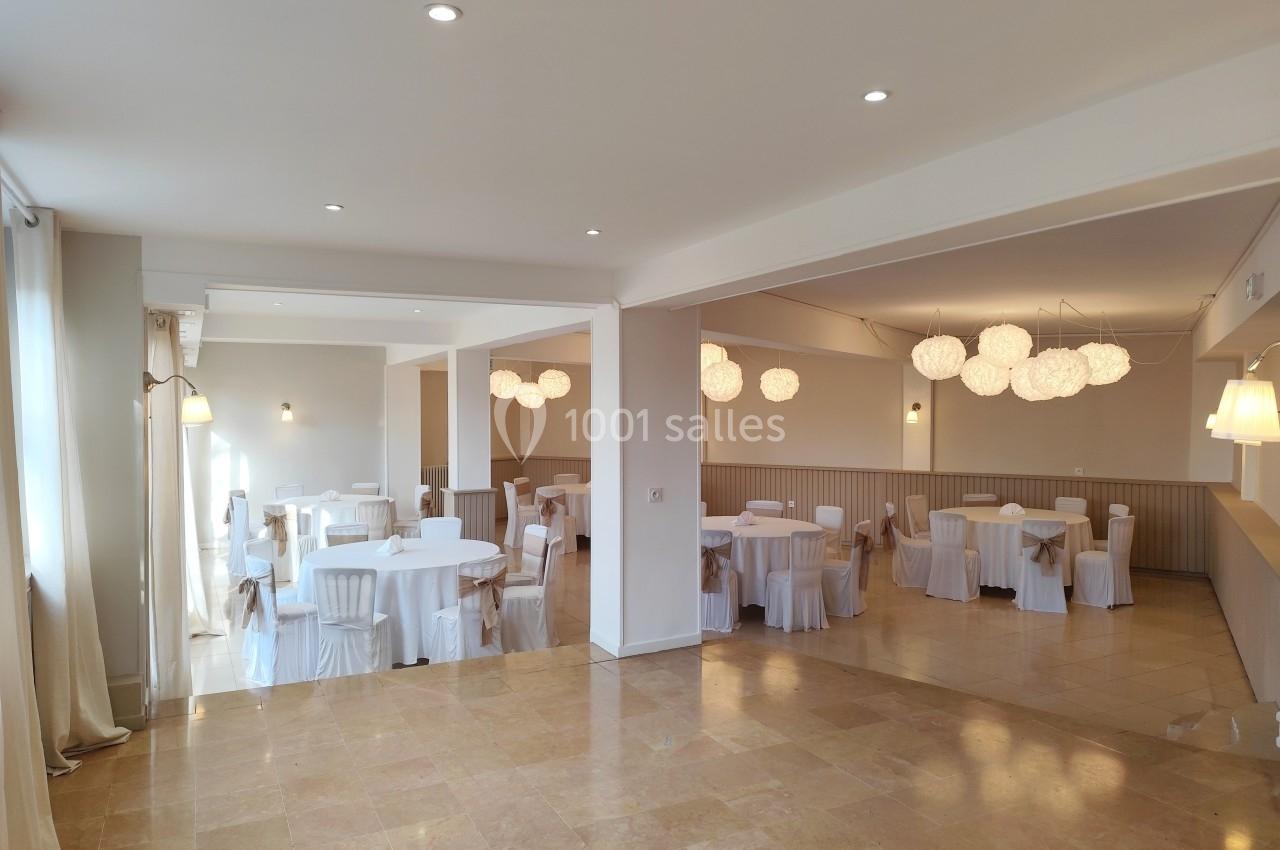 Salle lumineuse avec tables rondes dressées de nappes blanches, chaises assorties et suspensions décoratives modernes.