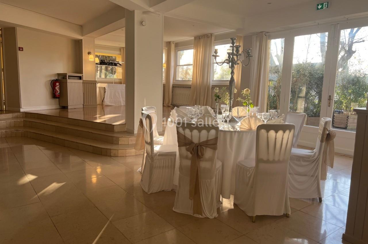 Salle lumineuse avec table ronde dressée pour un repas, nappes blanches et chaises décorées de rubans beiges.