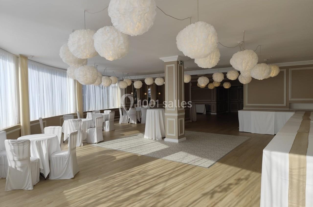 Salle lumineuse avec tables rondes et rectangulaires recouvertes de nappes blanches, décorée de suspensions en papier blanc.